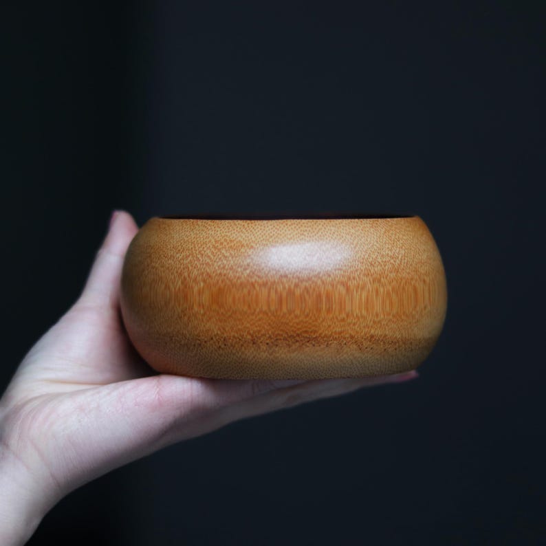 K&ouml;nnte beinhalten: Eine kleine, runde Holzschale mit nat&uuml;rlicher, hellbrauner Oberfl&auml;che. Die Schale wird in einer Hand vor einem dunklen Hintergrund gehalten. Die glatte, geschwungene Form der Schale und die Holzmaserung sind sichtbar.