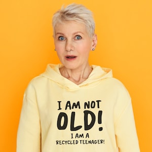 Könnte beinhalten: Eine Frau trägt einen gelben Hoodie mit dem Aufdruck "Ich bin nicht alt! Ich bin ein recycelter Teenager!" Sie schaut mit einem überraschten Ausdruck in die Kamera.