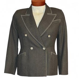 Pode incluir: Um blazer cinza carvão de abotoamento duplo com botões prateados e acabamentos cinza claro. O blazer tem lapelas entalhadas, dois bolsos frontais e um corte sob medida. O casaco é exibido em um manequim.