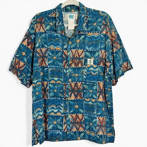 May include: Blue short-sleeve button-down shirt with a geometric pattern in shades of blue, orange, and red. The shirt has a collared neckline, wooden buttons, and a chest pocket. The tag reads "Duke Island".