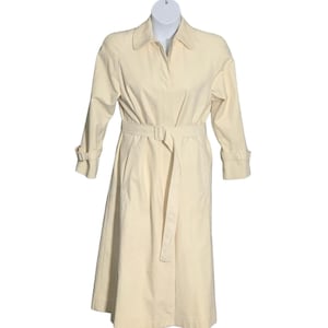 Puede incluir: Un abrigo tipo gabardina de color crema con cinturón en la cintura y mangas largas. El abrigo presenta un cuello clásico, puños abotonados y un cinturón a juego. El abrigo se muestra en un maniquí.