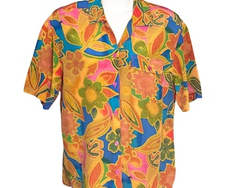 Könnte beinhalten: Ein kurzärmeliges Hemd mit einem auffälligen Blumenmuster. Das Hemd zeigt ein farbenfrohes Muster aus orangefarbenen, gelben, blauen und rosa Blumen und Blättern. Es hat eine Brusttasche und einen klassischen Kragen.