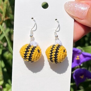 May include: A pair of handmade crochet bee earrings. The earrings feature yellow and black striped bodies with silver-colored hooks. The earrings are displayed on a white card against a background of green foliage and purple flowers.
