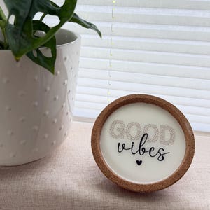 Könnte beinhalten: Eine runde Kerze mit einem Korkrand und einer weißen Wachsoberfläche, die den Schriftzug GOOD vibes in einer Kombination aus gepunkteter und kursiver schwarzer Schrift zeigt, ergänzt durch ein kleines schwarzes Herz unter dem Text.