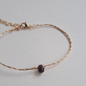 Könnte beinhalten: Ein zartes goldenes Armband mit einem gedrehten Drahtdesign und einer einzigen dunkelvioletten Perle.