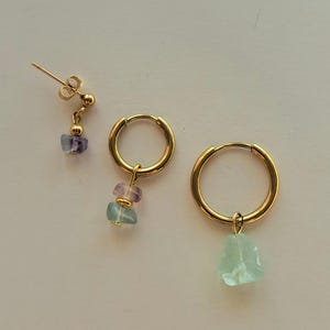 May include: A pair of gold hoop earrings with a single light blue gemstone dangling from one hoop and a stack of three gemstones, light blue, pink, and purple, dangling from the other hoop. The earrings are on a white background.
