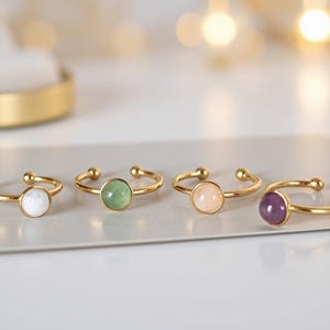 Könnte beinhalten: Vier goldfarbene, verstellbare Ringe mit unterschiedlich farbigen Edelsteinen: weiß, grün, rosa und lila. Jeder Ring hat einen kleinen, runden Edelstein in einem goldenen Band. Die Ringe werden auf einer weißen Oberfläche präsentiert.