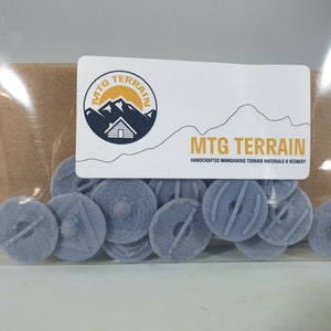 May include: A transparent bag containing multiple small, pale grey miniature shields. The shields feature embossed designs and are labelled with a white sticker that reads "MTG TERRAIN HANDCRAFTED WARGAMING TERRAIN MATERIALS & SCENERY".