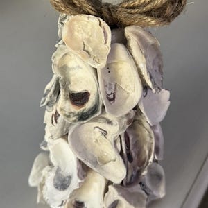 May include: A decorative bell-shaped ornament crafted from oyster shells. The shells are primarily off-white with subtle brown and gray markings. A natural twine bow adorns the top, adding a rustic touch. This coastal-themed item is suitable for home decor.
