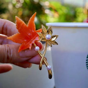 Peut inclure: Broche dorée représentant deux feuilles d'érable. L'une est orange, l'autre dorée avec des perles. La broche a une esthétique délicate inspirée de la nature, parfaite pour ajouter une touche d'élégance à n'importe quelle tenue.