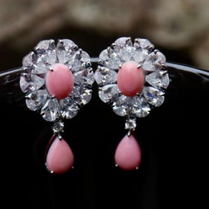 Può includere: Un paio di orecchini eleganti con una gemma rosa ovale centrale circondata da cristalli chiari e scintillanti in un design floreale. Ogni orecchino ha un pendente a goccia rosa. Sfondo scuro.