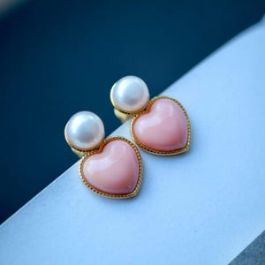 Può includere: Un paio di orecchini a forma di cuore dorati con accenti di perle rosa e bianche.