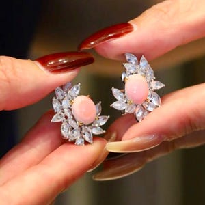Può includere: Un paio di orecchini eleganti con una gemma rosa ovale centrale, circondata da cristalli trasparenti disposti a motivo floreale. Gli orecchini sono mostrati da vicino, evidenziando il loro design intricato e i cristalli scintillanti.
