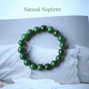 Könnte beinhalten: Ein Jade-Armband aus polierten, runden grünen Perlen. Das Armband wird auf einer weißen Oberfläche mit dem Text "Natural Nephrite" darüber gezeigt. Die Perlen haben eine tiefe grüne Farbe.