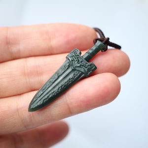 May include: Dark green pendant necklace featuring a detailed dagger design. The dagger has an ornate hilt and blade, with a textured pattern. The pendant is attached to a brown cord.