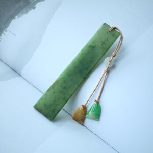 Op de afbeelding: Een rechthoekige jade bladwijzer met een lichtgroene tint en donkergroene vlekken. Het heeft een beige koord met twee kleine, klokvormige bedels, één goudkleurig en één groen. De bladwijzer ligt op een open boek.