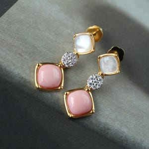 May include: A pair of gold drop earrings featuring a square pink stone at the base, a small circular cluster of clear stones in the middle, and a square white stone at the top, all set in a polished gold metal frame with a post back closure.