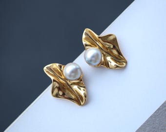 May include: A pair of gold-colored leaf-shaped earrings, each featuring a white pearl. The earrings have a textured, organic design, with the pearl centered on the leaf. The background is a mix of white, gray, and dark gray.