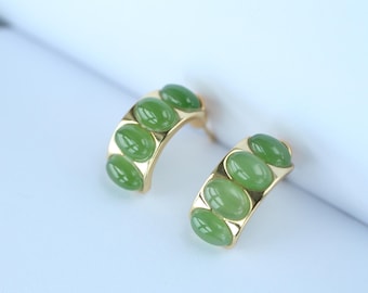 May include: A pair of gold-toned hoop earrings, each adorned with three oval, light green gemstones. The earrings have a curved design, with the gemstones set in a row. The background is a soft, blurred white.
