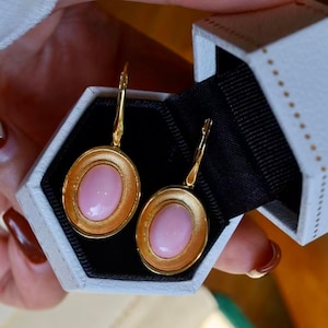 Può includere: Un paio di orecchini d'oro con pietre ovali rosa incastonate in una cornice d'oro. Gli orecchini sono in una scatola bianca con interno nero.