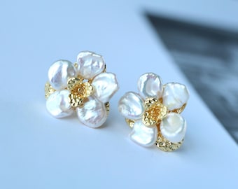 May include: A pair of floral-shaped earrings. Each earring features a cluster of white, irregularly shaped pearls arranged around a gold-colored center. The gold base has a textured, hammered appearance, creating a contrast with the smooth pearls.
