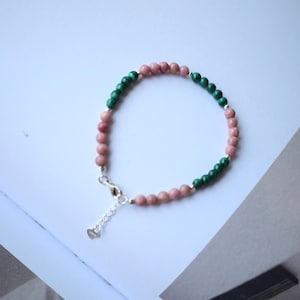 Op de afbeelding: Een armband met afwisselend roze en groene edelsteen kralen. De sluiting en verlengketting zijn zilverkleurig. De kralen zijn klein en rond, gelijkmatig verdeeld. De armband ligt op een witte ondergrond.