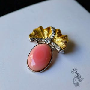 Peut inclure: Broche en or et argent avec une grosse pierre rose ovale. La broche présente un motif en forme d'éventail doré avec des accents argentés et de petites pierres précieuses transparentes. La pierre rose est sertie dans un cadre argenté.