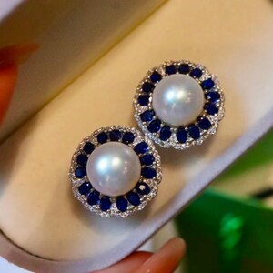 Op de afbeelding: Een paar parel oorknopjes. Elke oorbel heeft een grote, ronde, witte parel omgeven door een halo van blauwe edelstenen en kleinere heldere stenen, gezet in een zilverkleurig metaal. De oorbellen worden gepresenteerd op een lichtgekleurd oppervlak.