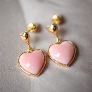 Può includere: Un paio di orecchini a forma di cuore dorati con pietre a forma di cuore rosa che pendono dalla parte inferiore.