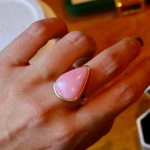 Può includere: Un anello con pietra preziosa rosa a forma di goccia con una fascia color oro. L'anello è indossato su un dito, mettendo in mostra la pietra lucida rosa chiaro. La fascia dorata crea un bordo a contrasto attorno alla pietra.