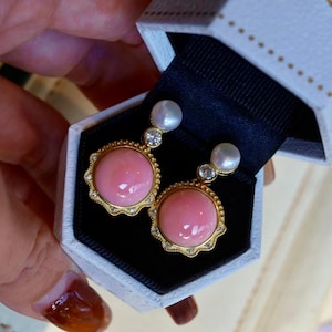Può includere: Un paio di orecchini in corallo rosa con montatura in oro e una perla a goccia. Gli orecchini sono in una scatola bianca con interno nero.
