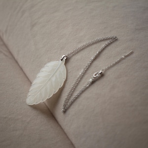 Puede incluir: Un delicado collar con un colgante de pluma blanca con detalles intrincados. El colgante cuelga de una fina cadena de plata. El collar se exhibe sobre un fondo de tela de tono neutro, resaltando el elegante diseño de la joya.