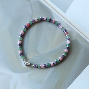 Pode incluir: Um bracelete multicolorido com contas quadradas verdes, rosa e roxas. O bracelete tem um fecho magnético.