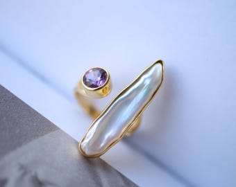 May include: A gold ring with a large white pearl and a small purple gemstone. The ring is designed to be worn on the knuckle.