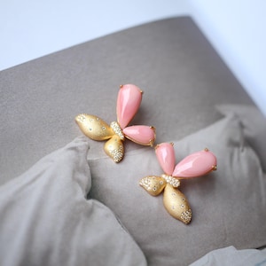 Può includere: Un paio di orecchini a farfalla dorati con accenti di pietre rosa. Gli orecchini sono delicati e femminili.