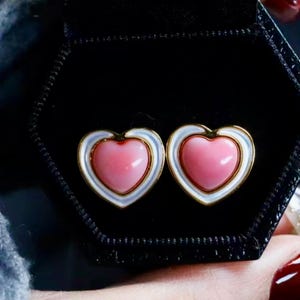 Può includere: Un paio di orecchini a forma di cuore con un centro rosa e un bordo bianco e dorato.