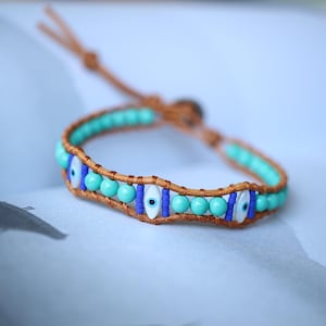 Puede incluir: Un brazalete de cuero marrón con cuentas turquesa y azules y dijes de ojo turco.