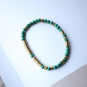 Op de afbeelding: Een armband met groene malachiet kralen en goudkleurige cilindrische kralen. De armband wordt gepresenteerd op een wit en grijs oppervlak, waarbij het ontwerp en het kleurcontrast worden benadrukt.