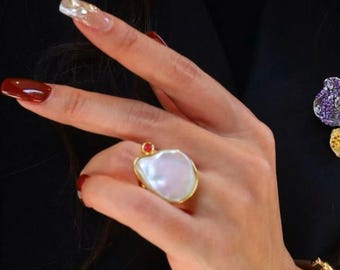 May include: A gold ring with a large, white, irregular-shaped pearl and a small red gemstone. The ring is on a person's finger.
