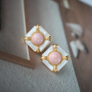 Può includere: Un paio di orecchini quadrati con montatura dorata. Ogni orecchino presenta un cerchio rosa centrale circondato da un quadrato bianco iridescente. La montatura dorata presenta dettagli decorativi. Gli orecchini sono su uno sfondo neutro.
