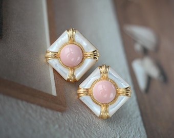 May include: A pair of square-shaped earrings with a gold-colored frame. Each earring features a central pink circle surrounded by a white, iridescent square. The gold frame has decorative detailing. The earrings are set against a neutral background.