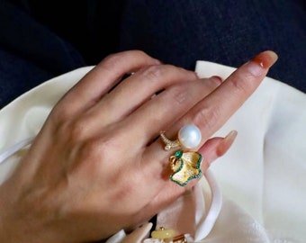 May include: A gold ring with a pearl and a green and gold leaf design. The ring is on a finger.