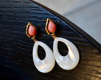 May include: A pair of gold-toned earrings with a teardrop design. Each earring features a coral-colored, teardrop-shaped stone set in a gold frame, with a larger, white, open teardrop pendant below. The earrings are displayed on a dark surface.