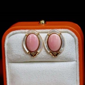 Può includere: Un paio di orecchini ovali con pietre di corallo rosa incastonate in oro e madreperla bianca. Gli orecchini sono esposti in una scatola di velluto rosso.