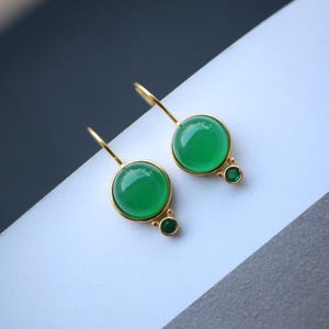 Peut inclure: Une paire de boucles d'oreilles pendantes dorées. Chaque boucle d'oreille présente une grosse pierre verte ronde et vibrante sertie dans une lunette dorée. Une petite pierre verte est sertie en dessous. Les boucles d'oreilles sont présentées sur une surface blanche.