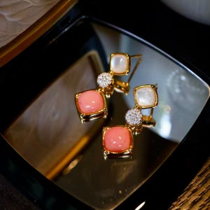 May include: A pair of gold drop earrings featuring a square white stone at the top, a small circular cluster of clear stones in the middle, and a larger square pink stone at the bottom. The earrings are displayed on a reflective surface.