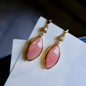 Può includere: Un paio di orecchini pendenti con una montatura dorata, ciascuno con una pietra rosa a forma ovale. Una piccola perla rotonda è sospesa sopra ogni pietra rosa. Gli orecchini sono presentati su una superficie bianca.