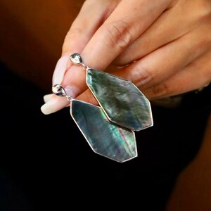 May include: A pair of silver-toned earrings with iridescent, dark green, and blue geometric pendants. The earrings are held up against a dark background, showcasing their unique shape and color.