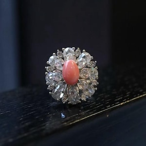 Può includere: Un anello d'argento con una pietra centrale ovale color corallo, circondata da numerose gemme chiare e scintillanti. L'anello è incastonato su uno sfondo scuro, evidenziando il suo design intricato e la brillantezza delle pietre.
