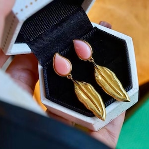 Può includere: Un paio di orecchini a forma di piuma dorati con pietre rosa a forma di goccia. Gli orecchini sono in una scatola bianca con interno nero.